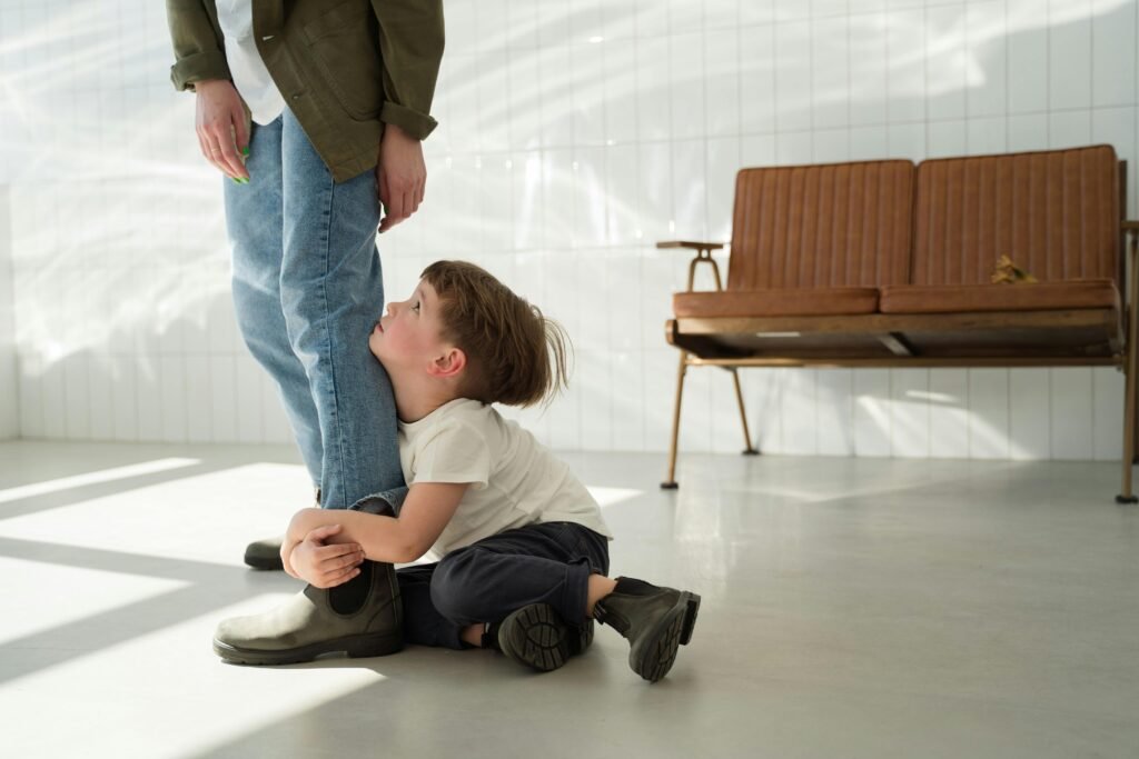A young boy clings to a parent's leg indoors, expressing emotion in natural light.