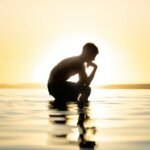 A man is silhouetted against a sunset while sitting on a lake, creating a reflective mood.